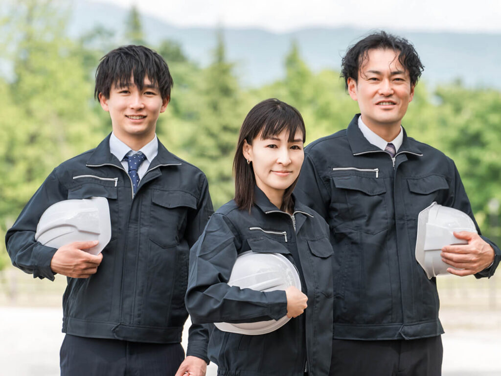 ヘルメットを手にした作業服姿の3人のスタッフが、笑顔で並んでいる